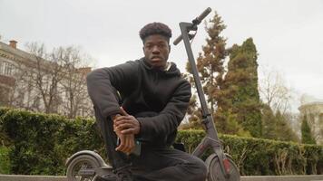 A young African-American man in a black sweater and black jeans and sneakers sits on an electric scooter near the lawn and looks at the camera with a smartphone in his hands. video