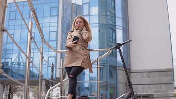 Young stylish blonde girl in a beige trench coat and jeans stands with an electric scooter with a modern high-rise on the background and takes a selfie video