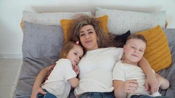 mamá y hija y hijo mentira en cama juntos abrazando mamá y niños son dormido y repentinamente abierto su ojos y Mira a el cámara. el familia es vestido en el mismo básico blanco camisetas video