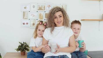 un' ragazzo, un' ragazza e un' madre siamo seduta a un' tavolo nel un' elegante bianca moderno cucina nel scandinavo design e guardare a il telecamera. famiglia nel bianca magliette e jeans video