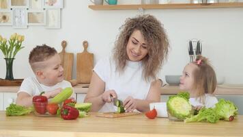 Jeune blond femme avec frisé cheveux coupes des légumes et communique avec les enfants séance à une table dans le cuisine dans une scandinave conception hygge. fermer vue de une couteau dans le mains video