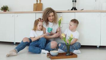 un chico, un niña y un madre sentar en el piso en un elegante blanco moderno cocina y tener desayuno con cereales. familia en blanco camisetas y pantalones video