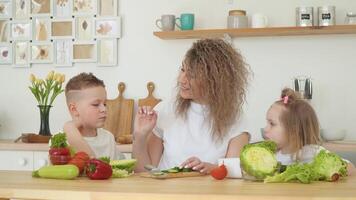 jovem Loiras mulher com encaracolado cabelo dá uma gosto do uma fatia do pepino para dela filho e filha sentado às uma mesa dentro a cozinha. a menina recusa para comer legumes video