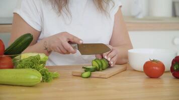 Young blonde woman with curly hair cuts a cucumber into rings while sitting at a table in the kitchen in a Scandinavian design hygge. Close-up view of hand movements. Healthy food and diet concept video