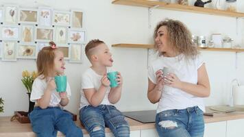 garoto, menina e mãe Piada e beber uma beber a partir de azul canecas sentado em a mesa dentro uma à moda branco moderno cozinha dentro escandinavo Projeto. família dentro branco Camisetas e jeans video