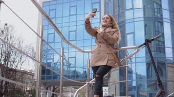 jung stilvoll blond Mädchen im ein Beige Graben Mantel und Jeans steht mit ein elektrisch Roller mit ein modern Hochhaus auf das Hintergrund und nimmt ein Selfie video