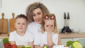 kinderen met mam zittend Bij een tafel in een helder keuken. vers groenten zijn Aan de tafel. gezond aan het eten. kinderen menu. de familie is gekleed in de dezelfde eenvoudig kleren van wit en blauw kleuren video