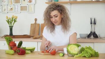 Young blonde woman with curly hair slices a red tomato sitting at a table in the kitchen in a Scandinavian Hygge design. Healthy food and diet concept video