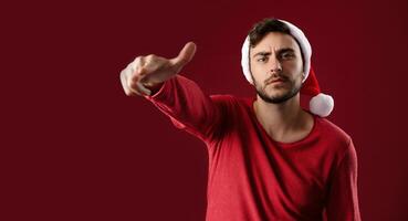 Young handsome caucasian guy in a red sweater and Santa hats stands on red background in studio and Shows a finger at the camera. photo