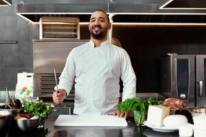 Handsome young African chef standing in professional kitchen in restaurant preparing a meal of meat and cheese vegetables. photo