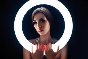 A beautiful young Caucasian girl with a beautiful make-up looks out from behind a circular lamp for portrait photo shooting