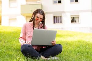 Photo of european woman 25s sitting on green grass in park with legs crossed during summer day while using laptop. Caucasian female hipster with dreadlocks and pink sunglasses on use laptop