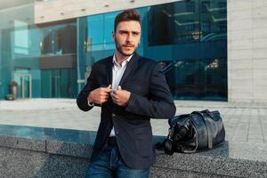 hermoso joven empresario con un barba y en un negocio traje en pie en el calle en contra el antecedentes de el oficina edificio siguiente a un cómodo elegante cuero bolsa. foto