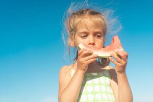 Funny little girl eat watermelon sunny summer day at ocean beach. Cute caucasian female child enjoy summer fruit bite slice of watermelon. Happy childhood. photo