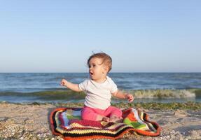 contento bebé sonriente y ondulación mano, sentado en blanco arenoso tropical playa en el alfombra foto