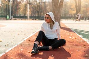 Outdoor portrait of young beautiful woman with long in sunglasses and a white hooded sweater sitting on the sportsground track photo