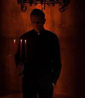 Young Catholic Praying priest. Portrait of priest Next to the candles prays with his hands folded near the face. Red wall on the background. Indoor handsome portrait of religion man photo