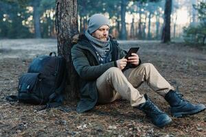 adult handsome Male traveler Sits On autumn pine forest Near tree, Holding At Tablet Recreation Resting Travel Destination Concept photo