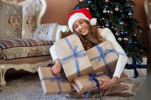 Navidad. mujer vestido blanco suéter Papa Noel sombrero y pantalones sentado en el piso cerca Navidad árbol con presente caja foto