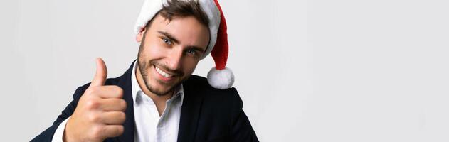 joven hermoso caucásico chico en negocio traje y Papa Noel sombreros soportes en blanco antecedentes en estudio carita y demostración pulgares arriba. foto