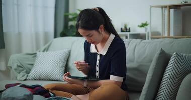 imágenes lento movimiento disparo, asiático adolescente mujer sentado en sofá participación crédito tarjeta haciendo en línea pago en digital tableta, preparación viaje maleta a hogar. video