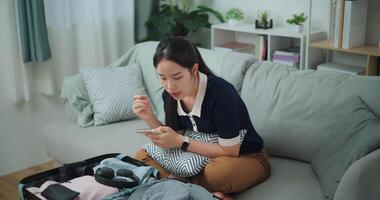 Footage high angle view shot, Asian teenager woman sitting on sofa making checklist of things to pack for travel, Preparation travel suitcase at home. video