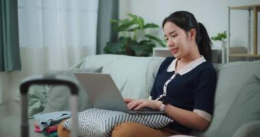 Footage dolly shot, Asian teenager woman sitting on sofa using laptop for prepare booking hotel and airplane ticket for travel. backpacker travel concept. video