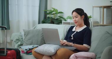 metraggio dolly sparo, asiatico adolescente donna seduta su divano utilizzando il computer portatile per preparare prenotazione Hotel e aereo biglietto per viaggio. zaino in spalla viaggio concetto. video