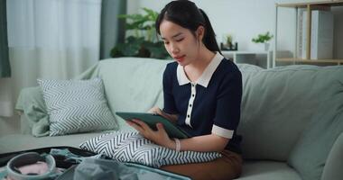 métrage Zoom dans tir, asiatique adolescent femme séance sur canapé en utilisant numérique tablette fabrication vérifier liste de des choses à pack pour voyage, préparation Voyage valise à maison. video