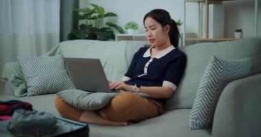 antal fot långsam rörelse skott, asiatisk tonåring kvinna Sammanträde på soffa använder sig av bärbar dator för förbereda bokning hotell och flygplan biljett för resa. backpacker resa begrepp. video