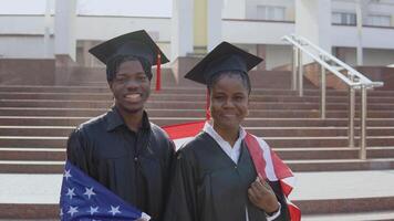 afrikanisch amerikanisch Mann und Frau Stand Seite durch Seite gegenüber das Kamera im schwarz Roben und Platz Hüte von Absolvent Studenten mit das USA Flagge auf ihr Schultern video