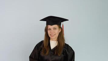 A young caucasian female student in a black robe touches a square hat . White background video