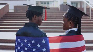 African American man and woman standing side by side with their backs to the camera. They have the British flag on their shoulders. A University building on background video