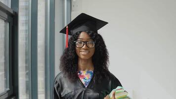 Young African American female university graduate standing with textbooks in hands near a large modern window with black frames. Female student wearing a black robe, square master's hat and glasses video