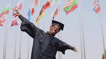 muito jovem africano americano masculino graduado em pé dentro frente do a Câmera com uma diploma dentro dele mãos. a aluna chapéu e carrinhos lado de fora com a internacional bandeiras em fundo. video