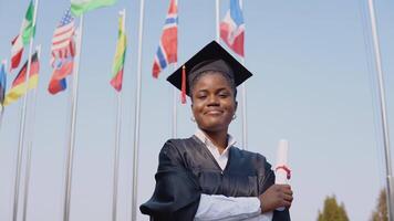 jovem sorridente africano americano fêmea graduado em pé dentro frente do a Câmera com uma diploma dentro dela mãos. a aluna carrinhos lado de fora com a internacional bandeiras em fundo. video