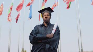 joven africano americano masculino graduado en pie en frente de el cámara con un diploma en su manos. el estudiante tiene un cuadrado maestría sombrero y soportes fuera de con el internacional banderas en antecedentes. video