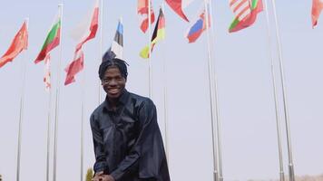 Young african american male graduate expresses happiness and joy, while standing in front of the camera with a diploma s in his hands. The stands outside with the international flags on background. video