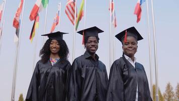 drei Afroamerikaner Universität Absolventen Stand draußen gegen das Hintergrund von Flaggen von um das Welt. Studenten sind gekleidet im das festlich Uniform von das Meister video