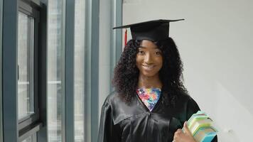 Young African American female university graduate standing with textbooks in hands near a large modern window with black frames. video