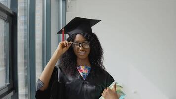 Young African American female university graduate standing with textbooks in hands near a large modern window with black frames. Female student wearing a black robe, square master's hat and glasses video