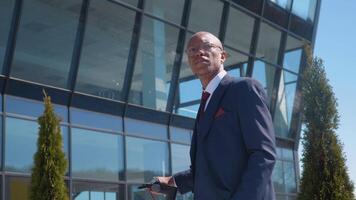 A young business man in a classic blue business suit stands with an electric scooter on the street near a modern mirror building. Portrait to the waist video