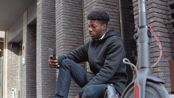en ung afrikansk amerikan man i en svart Tröja och svart jeans och gymnastikskor sitter på de trottoar nära en byggnad med en mörk tegel Fasad. gadgetar för ett aktiva livsstil och kommunikation video