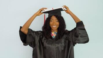 A young African American woman in a black gown and a master's hat stands upright holding a diploma over her hat and looks into the camera. Portrait of half height video