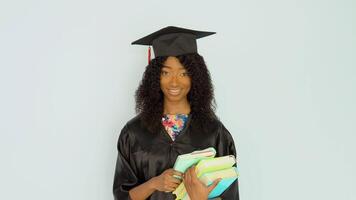 A young African American woman in a black robe and a master's hat stands straight holding textbooks. She looks at the camera. Portrait of half height video