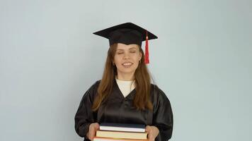 A young Caucasian woman in a black robe and a master's hat stands straight holding textbooks and looks at the camera. Portrait of half height video