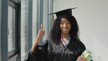Young African American female university graduate stands with textbooks in one hand and shows gesture good with big finger up near a large modern window with black frames. video