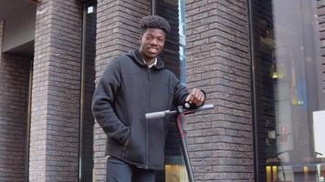 een jong Afrikaanse Amerikaans Mens in gewoontjes donker kleren met een scooter staat in de buurt een gebouw met een donker steen facade video