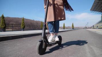 Young girl student in jeans and a coat rides on an electric scooter. Close up view of the scooter and wheels from front. Ecological city transport video