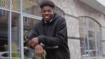 A young African-American man in a black sweater and black jeans and sneakers leaned on an electric scooter near a building with large windows and looked at the camera with a smartphone in his hands. video